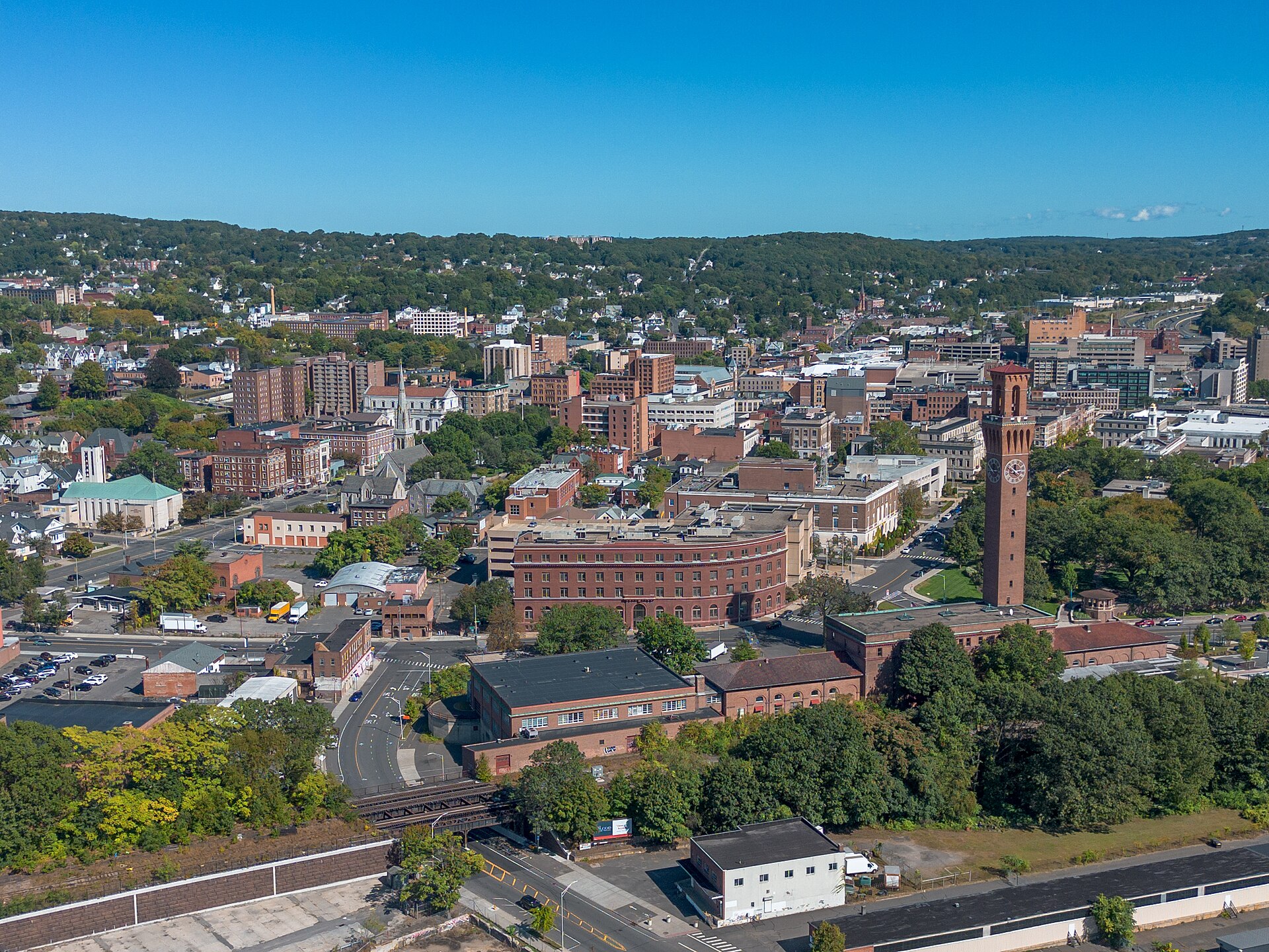 Downtown Waterbury’s brick buildings and the Union Station clock tower among green hills, seen from above on a clear day.