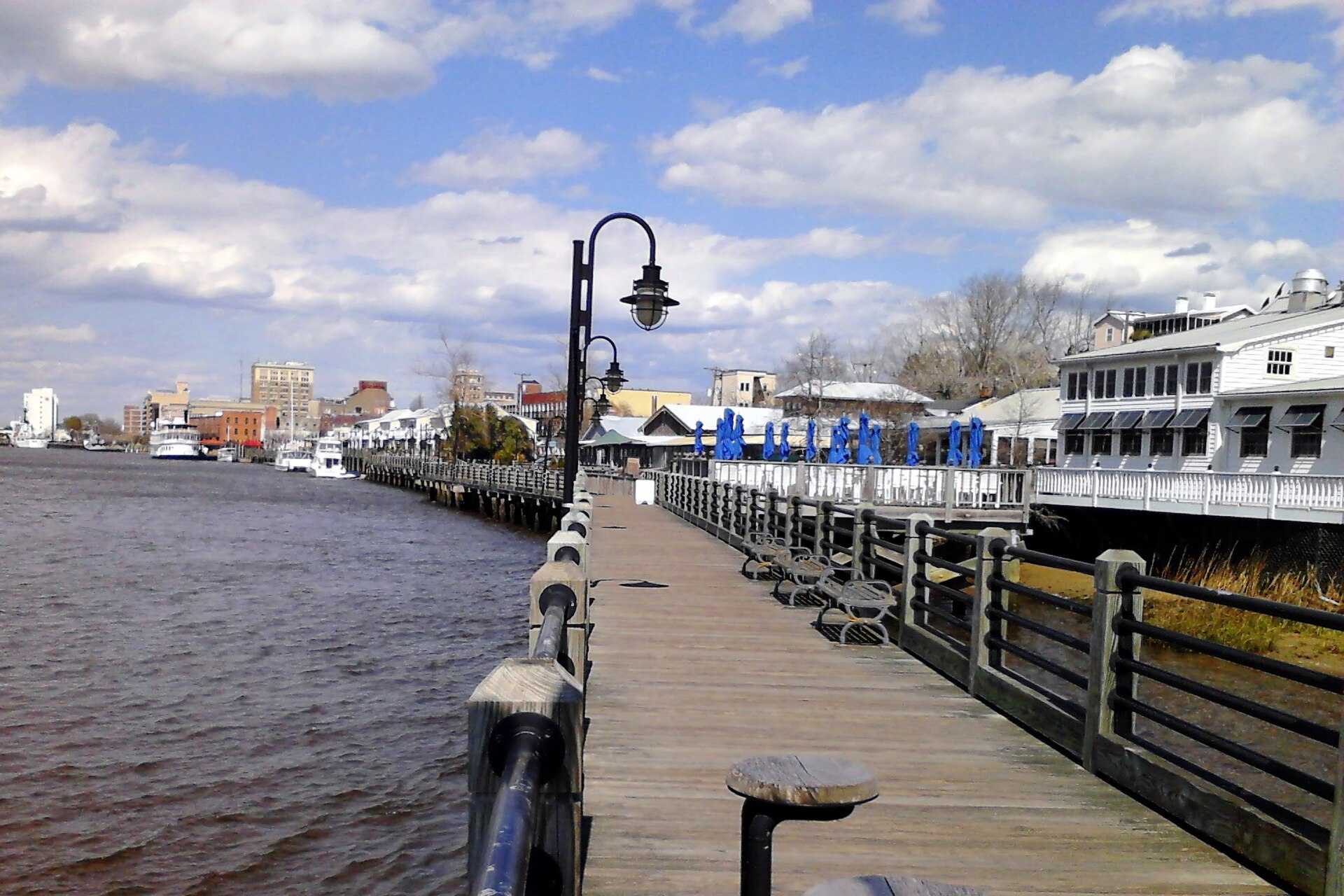 The Riverwalk's wooden boardwalk along the Cape Fear River, with downtown Wilmington beyond, under a partly cloudy sky.